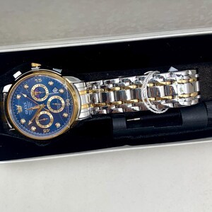 K&ouml;nnte beinhalten: Eine gold- und silberfarbene Armbanduhr mit blauem Zifferblatt, pr&auml;sentiert in einer wei&szlig;en Box mit schwarzem Innenraum. Die Uhr verf&uuml;gt &uuml;ber mehrere Hilfszifferbl&auml;tter und eine Datumsanzeige. Das Armband ist eine Kombination aus goldenen und silbernen Gliedern. Ein Uhren-Einstellwerkzeug ist enthalten.