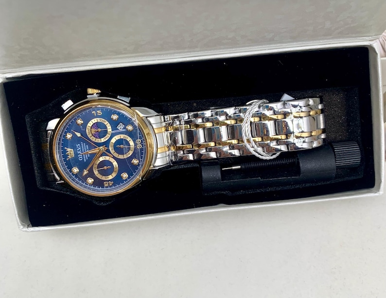 K&ouml;nnte beinhalten: Eine gold- und silberfarbene Armbanduhr mit blauem Zifferblatt, pr&auml;sentiert in einer wei&szlig;en Box mit schwarzem Innenraum. Die Uhr verf&uuml;gt &uuml;ber mehrere Hilfszifferbl&auml;tter und eine Datumsanzeige. Das Armband ist eine Kombination aus goldenen und silbernen Gliedern. Ein Uhren-Einstellwerkzeug ist enthalten.