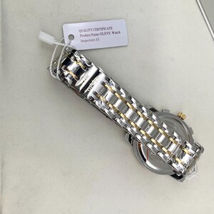 K&ouml;nnte beinhalten: Eine silber- und goldfarbene Armbanduhr mit Metallarmband. Das Zifferblatt ist sichtbar, und ein am Armband befestigtes Etikett lautet "QUALITY CERTIFICATE Product Name: OLEVS Watch Inspectors: 15."