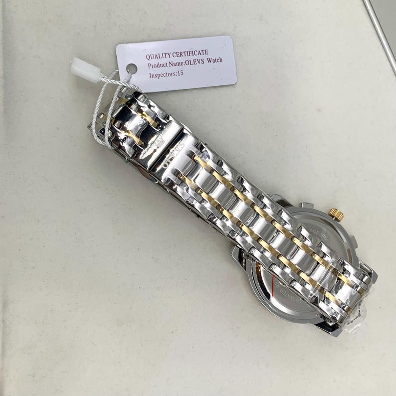 K&ouml;nnte beinhalten: Eine silber- und goldfarbene Armbanduhr mit Metallarmband. Das Zifferblatt ist sichtbar, und ein am Armband befestigtes Etikett lautet "QUALITY CERTIFICATE Product Name: OLEVS Watch Inspectors: 15."
