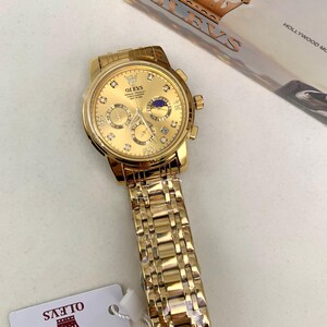 K&ouml;nnte beinhalten: Eine goldfarbene OLEVS Armbanduhr mit passendem Armband. Das Zifferblatt der Uhr zeigt ein Kronenlogo, mehrere Hilfszifferbl&auml;tter und eine Mondphasenanzeige. Die Uhr hat eine strukturierte, gl&auml;nzende Oberfl&auml;che und ist mit kleinen, funkelnden Akzenten versehen.