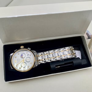 K&ouml;nnte beinhalten: Eine silber- und goldfarbene Armbanduhr mit wei&szlig;em Zifferblatt und mehreren Hilfszifferbl&auml;ttern, pr&auml;sentiert in einer wei&szlig;en Box mit schwarzem Innenraum. Die Uhr verf&uuml;gt &uuml;ber ein silber- und goldfarbenes Gliederarmband und ein Werkzeug zum Einstellen des Armbands.
