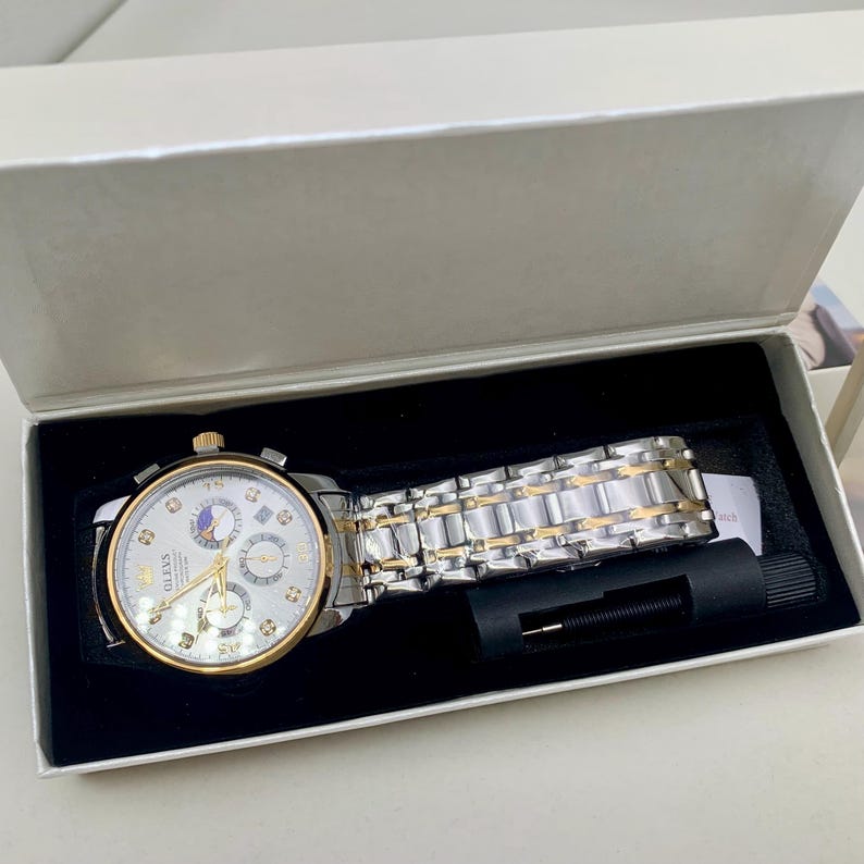 K&ouml;nnte beinhalten: Eine silber- und goldfarbene Armbanduhr mit wei&szlig;em Zifferblatt und mehreren Hilfszifferbl&auml;ttern, pr&auml;sentiert in einer wei&szlig;en Box mit schwarzem Innenraum. Die Uhr verf&uuml;gt &uuml;ber ein silber- und goldfarbenes Gliederarmband und ein Werkzeug zum Einstellen des Armbands.
