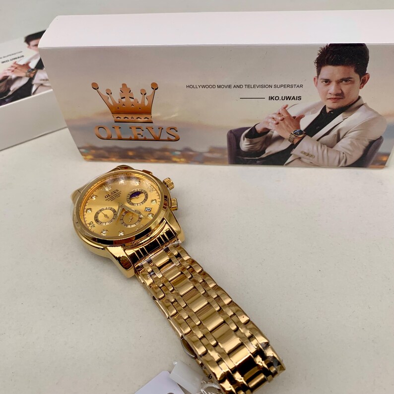 K&ouml;nnte beinhalten: Eine goldfarbene OLEVS Armbanduhr mit passendem Armband. Das Zifferblatt der Uhr verf&uuml;gt &uuml;ber mehrere Hilfszifferbl&auml;tter und eine Mondphasenanzeige. Die Uhr wird mit ihrer Box pr&auml;sentiert, die den Text "HOLLYWOOD MOVIE AND TELEVISION SUPERSTAR" und den Namen "IKO.UWAIS" tr&auml;gt.