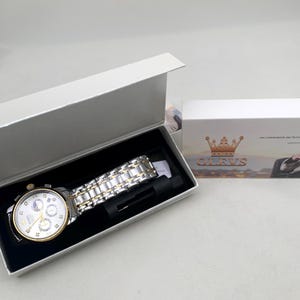 K&ouml;nnte beinhalten: Eine silber- und goldfarbene OLEVS-Uhr mit wei&szlig;em Zifferblatt, pr&auml;sentiert in einer offenen silbernen Schachtel. Die Uhr verf&uuml;gt &uuml;ber mehrere Hilfszifferbl&auml;tter und eine Datumsanzeige. Das Armband ist eine Kombination aus silbernen und goldfarbenen Gliedern. Die Schachtel befindet sich neben einer wei&szlig;en Schachtel mit dem OLEVS-Logo.