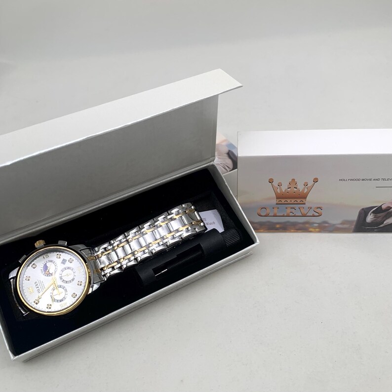 K&ouml;nnte beinhalten: Eine silber- und goldfarbene OLEVS-Uhr mit wei&szlig;em Zifferblatt, pr&auml;sentiert in einer offenen silbernen Schachtel. Die Uhr verf&uuml;gt &uuml;ber mehrere Hilfszifferbl&auml;tter und eine Datumsanzeige. Das Armband ist eine Kombination aus silbernen und goldfarbenen Gliedern. Die Schachtel befindet sich neben einer wei&szlig;en Schachtel mit dem OLEVS-Logo.