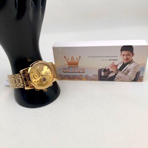 K&ouml;nnte beinhalten: Eine goldfarbene OLEVS-Armbanduhr mit passendem Armband wird auf einer schwarzen Mannequin-Hand gezeigt. Das Zifferblatt der Uhr hat mehrere Zifferbl&auml;tter und eine Mondphasenanzeige. Im Hintergrund befindet sich eine Schachtel mit dem OLEVS-Logo und einem Prominentenbild.