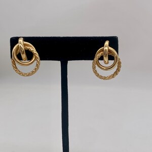 May include: A pair of gold-toned earrings with a double circle design. The outer circle is a braided rope design and the inner circle is a smooth, solid circle.