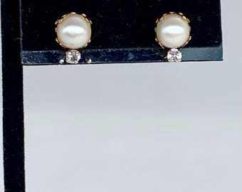 May include: A pair of gold-toned stud earrings. Each earring features a large, oval, off-white pearl surrounded by a decorative gold setting. A small, clear, round gemstone is set below the pearl. The earrings are displayed on a black surface.