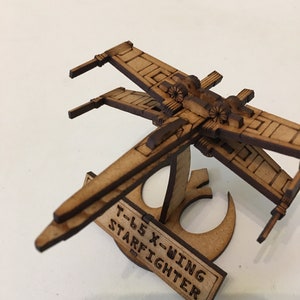Puede incluir: Un modelo de madera de un caza estelar X-Wing T-65 de Star Wars. El modelo está en un soporte con el símbolo de la Alianza Rebelde.