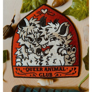 May include: Embroidered patch with two cartoon animals, embracing. The patch has a red border and the text "QUEER ANIMAL CLUB". The animals are white and grey with black accents. The patch is on a wooden surface.