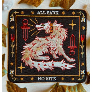 May include: Embroidered patch with a black background featuring a wolf with the text "ALL BARK" above and "NO BITE" below. The wolf is beige with red accents, and the design includes two red swords and floral elements.