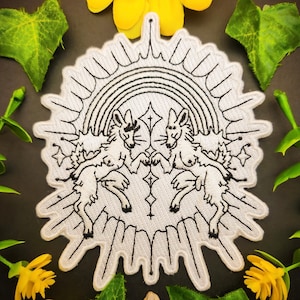 May include: White embroidered patch featuring two mythical creatures beneath a rainbow. The patch has a sunburst design around the edges, with black line art detailing. The patch is surrounded by green leaves and yellow flowers.