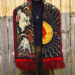 May include: Black scarf with red fringe and detailed illustrations. One side features a rabbit and bird with floral accents, the other a sun design. The scarf has a red border and is made of a soft material.