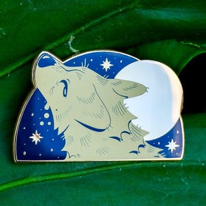 Könnte beinhalten: Emaille-Anstecknadel mit einem heulenden Wolf vor einem Vollmond in einem Nachthimmel. Die Anstecknadel hat einen goldenen Rand, einen blauen Hintergrund mit weißen Sternen und einen cremefarbenen Wolf. Ein einzigartiges Accessoire für Jacken, Taschen oder Hüte.