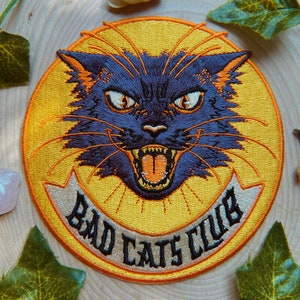 Könnte beinhalten: Gestickter Aufnäher mit einem wütenden schwarzen Kater mit orangefarbenen Akzenten auf gelbem Hintergrund. Der Aufnäher trägt den Schriftzug "BAD CATS CLUB" auf einem weißen Banner, ideal für Jacken oder Taschen.
