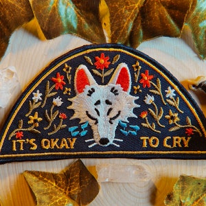May include: Embroidered patch with a navy blue background, featuring a white wolf face with red ears and floral accents. The patch reads "IT'S OKAY TO CRY" in white text, with a gold border.