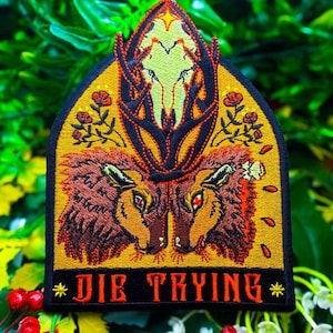 May include: An embroidered patch with a pointed arch shape featuring two brown deer heads facing each other, a central deer skull with antlers, and small red flowers on a yellow background. The bottom black border displays the text DIE TRYING.