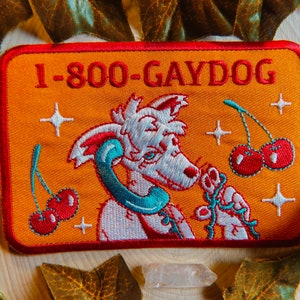 May include: Embroidered patch with the text "1-800-GAYDOG" on an orange background. The patch features a cartoon dog with a phone receiver, cherries, and star accents. The patch has a red border.