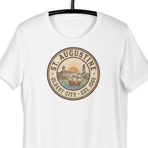 Op de afbeelding: Een wit T-shirt met korte mouwen en een cirkelvormige afbeelding op de borst. Het ontwerp toont een zeilschip, een vuurtoren en een fort met de tekst ST. AUGUSTINE OLDEST CITY - EST. 1565 in een bruin en beige kleurenpalet op het shirt.