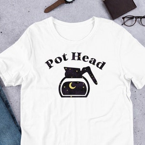 May include: White t-shirt featuring the words "Pot Head" above a coffee pot graphic with a night sky design. The coffee pot graphic includes a crescent moon and stars. The shirt is shown on a flat surface with jeans and accessories.
