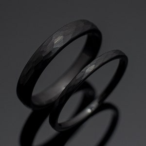 May include: Two black hammered wedding bands. The bands are made of a smooth, matte black metal. The bands are stacked on top of each other.