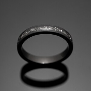 Peut inclure: Une bague gris fonc&eacute; avec une bande de taches argent&eacute;es. La bague est pos&eacute;e sur un fond sombre, avec son reflet visible en dessous. La bague est un bijou.