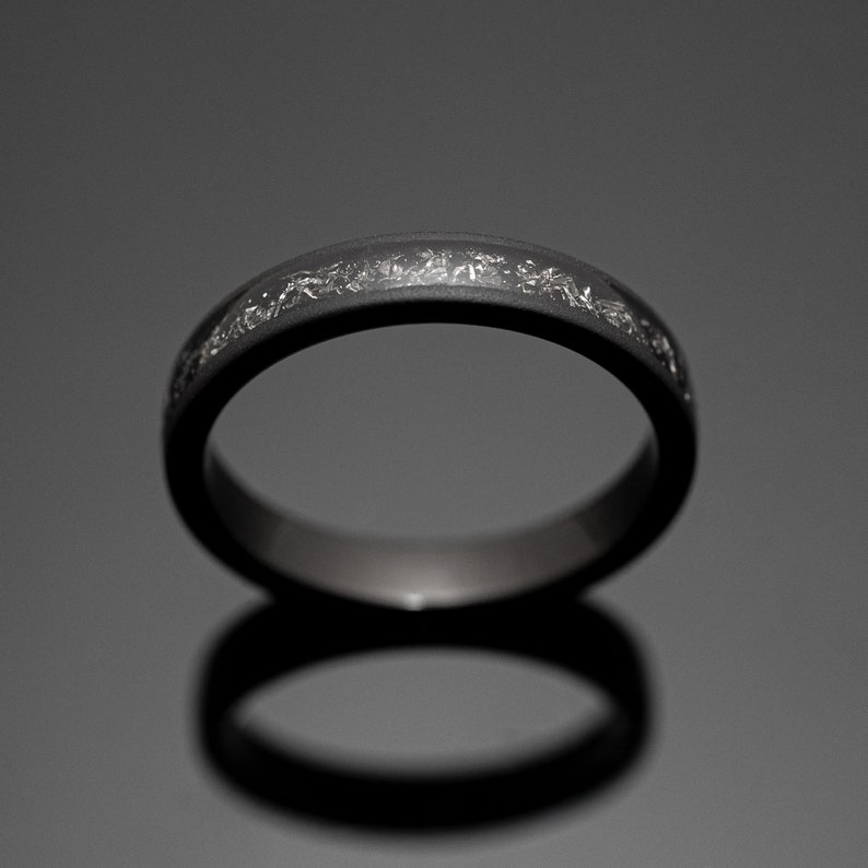 Peut inclure: Une bague gris fonc&eacute; avec une bande de taches argent&eacute;es. La bague est pos&eacute;e sur un fond sombre, avec son reflet visible en dessous. La bague est un bijou.