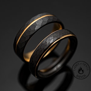 May include: Two black wedding bands with a hammered texture and a thin gold stripe. The rings are set against a dark background, with a logo that reads "Ladies & Gents Bands."