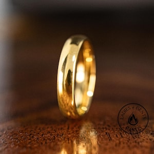 May include: A polished gold wedding ring. The ring is set against a dark wooden surface. The band has a classic, rounded design. The logo "Ladies & Gents Bands" is visible in the lower right corner. The ring's design is simple and elegant.