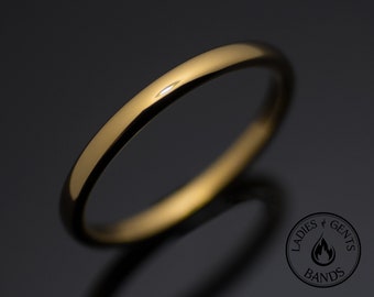 May include: A polished gold-tone band ring with a smooth, rounded surface and a reflective finish. The ring is positioned at an angle, showcasing its simple, circular design. A circular logo with the text LADIES GENTS BANDS is visible.