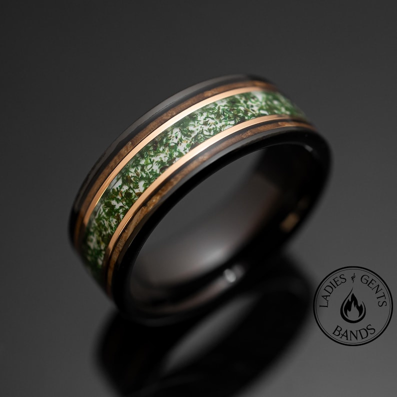 May include: A black ring featuring a central inlay of green and white moss-like material bordered by two thin rose gold bands. The ring displays a logo in the bottom right corner with the text LADIES GENTS BANDS and a flame icon.