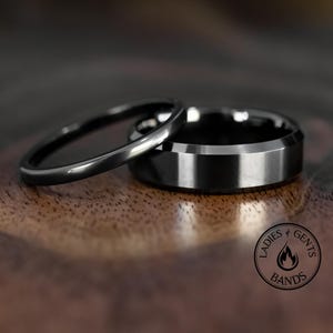 May include: Two black wedding rings. One ring is thin and rounded, the other is wider with a flat, beveled edge. The rings are displayed on a wooden surface. The image includes a circular logo with the text "Ladies & Gents Bands".