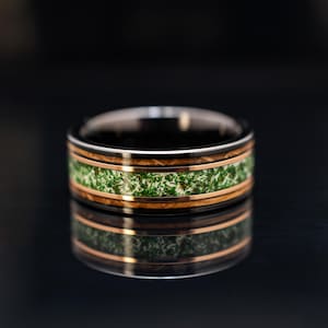 May include: A ring featuring a dark metallic band with a central inlay of green moss-like material, bordered by two thin rose gold-colored stripes and two outer bands of dark wood grain, creating a layered and textured appearance.