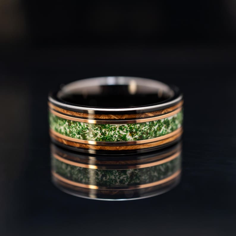May include: A ring featuring a dark metallic band with a central inlay of green moss-like material, bordered by two thin rose gold-colored stripes and two outer bands of dark wood grain, creating a layered and textured appearance.