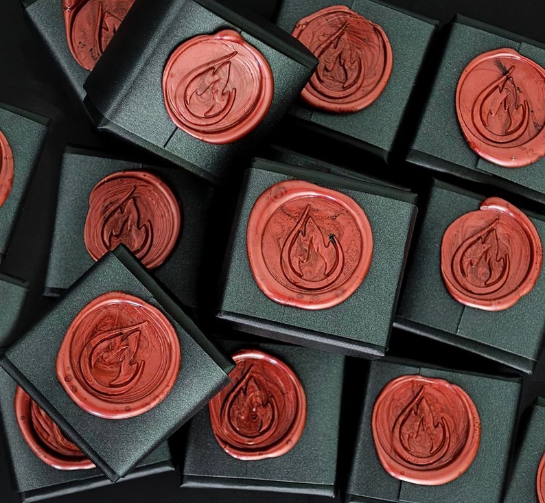 May include: An overhead shot shows several black gift boxes, each sealed with a reddish-brown wax seal. The seals feature a stylized flame design. The boxes are arranged closely together against a dark background, creating a pattern.