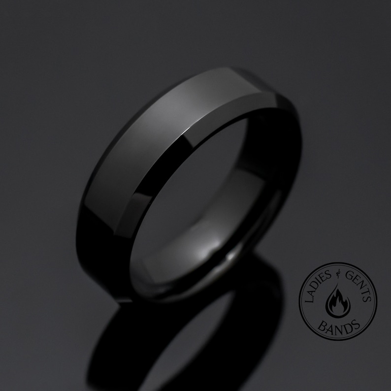 May include: A polished black ring with beveled edges and a smooth, reflective surface. In the bottom right corner, a circular logo features the text "LADIES & GENTS BANDS" surrounding a flame icon, indicating the brand identity of the jewelry.