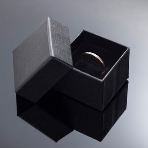 May include: A thin, polished rose gold band ring rests inside a square, black textured jewellery box. The box has a matching lid that is partially open, revealing the dark interior lining where the ring is positioned upright in the centre.