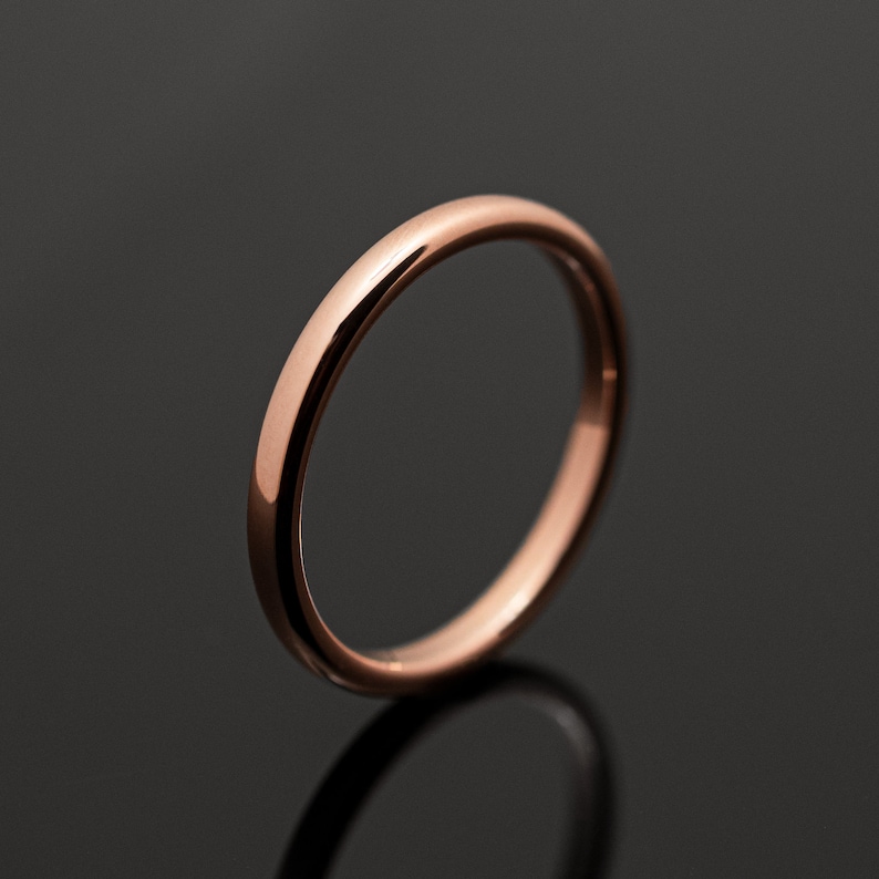 May include: A simple, polished rose gold band ring with a smooth, rounded profile. The metal surface has a warm, metallic pink hue and a reflective finish that catches the light, creating a clean and uniform appearance throughout the entire band.