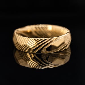May include: A gold-colored ring with a textured surface. The ring's design features a pattern of diagonal lines and curves, creating a unique visual texture. The ring is displayed on a reflective black surface.