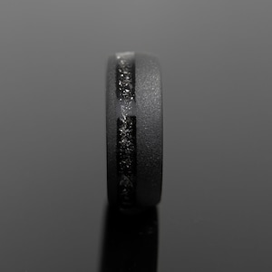 May include: A black ring with a textured surface and a central inlay of sparkling, dark material. The ring is shown in a side view, highlighting its sleek, modern design. The ring's width is approximately 0.3 inches.