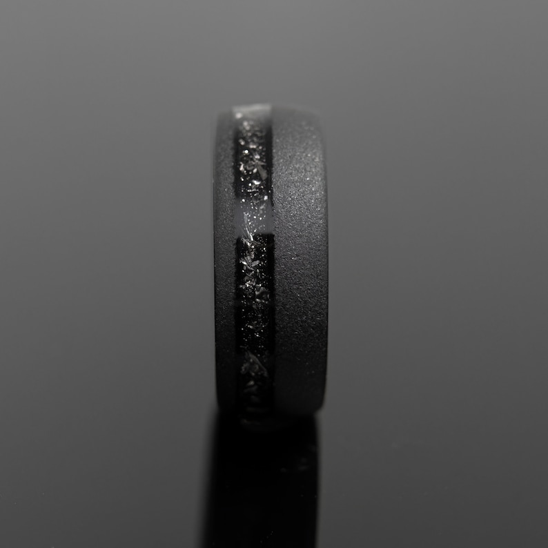 May include: A black ring with a textured surface and a central inlay of sparkling, dark material. The ring is shown in a side view, highlighting its sleek, modern design. The ring's width is approximately 0.3 inches.