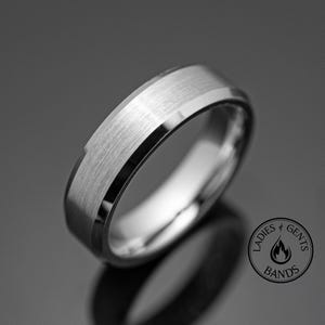 Könnte beinhalten: Ein silberfarbener Ring mit gebürsteter Mittelband und polierten Kanten. Der Ring hat ein flaches Profil und ein modernes Design. Das Bild enthält ein Logo mit der Aufschrift "Ladies Gents Bands".