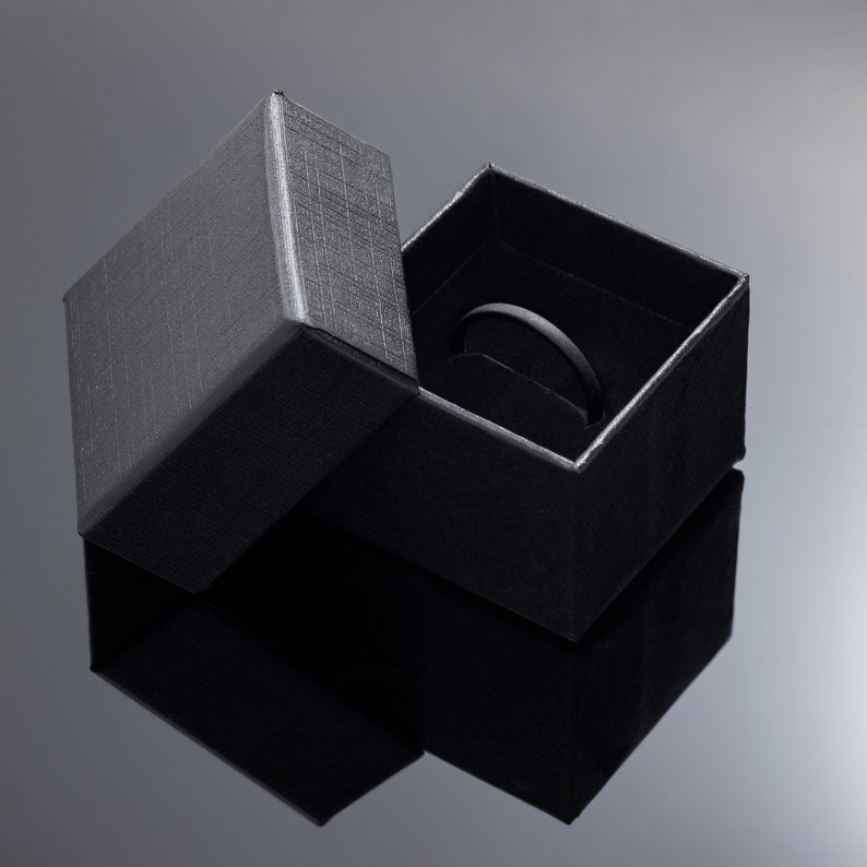 May include: An open black ring box with a textured exterior. The box is on a reflective surface, with the lid slightly ajar, revealing a ring holder inside. The box is designed to hold a ring.