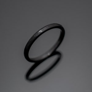 Op de afbeelding: Een donkergrijze gehamerde metalen ring. De ring heeft een getextureerd oppervlak en een eenvoudig, elegant ontwerp. De ring wordt getoond op een reflecterend oppervlak.