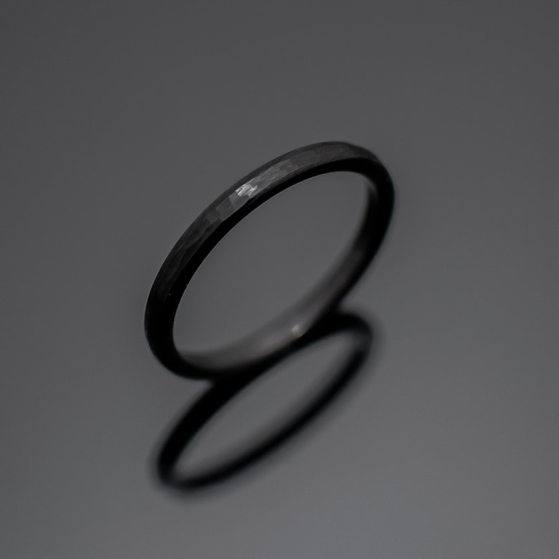 Op de afbeelding: Een donkergrijze gehamerde metalen ring. De ring heeft een getextureerd oppervlak en een eenvoudig, elegant ontwerp. De ring wordt getoond op een reflecterend oppervlak.