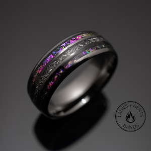 May include: A black ring with a band of colorful iridescent material. The ring has a dark, metallic finish and a textured design along the edges. The iridescent band displays shades of pink, purple, and green.