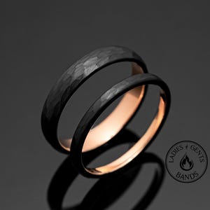 May include: Two black wedding bands with a hammered texture on the outside and a rose gold interior. The rings are displayed on a reflective surface, with a logo in the bottom right corner that reads "Ladies & Gents Bands."