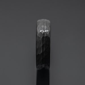 Op de afbeelding: Een donkergrijze ring met een gehamerde textuur. De ring heeft een bandbreedte van ongeveer 6 mm. Het oppervlak van de ring reflecteert licht, waardoor highlights en schaduwen ontstaan. De ring is een sieraad.