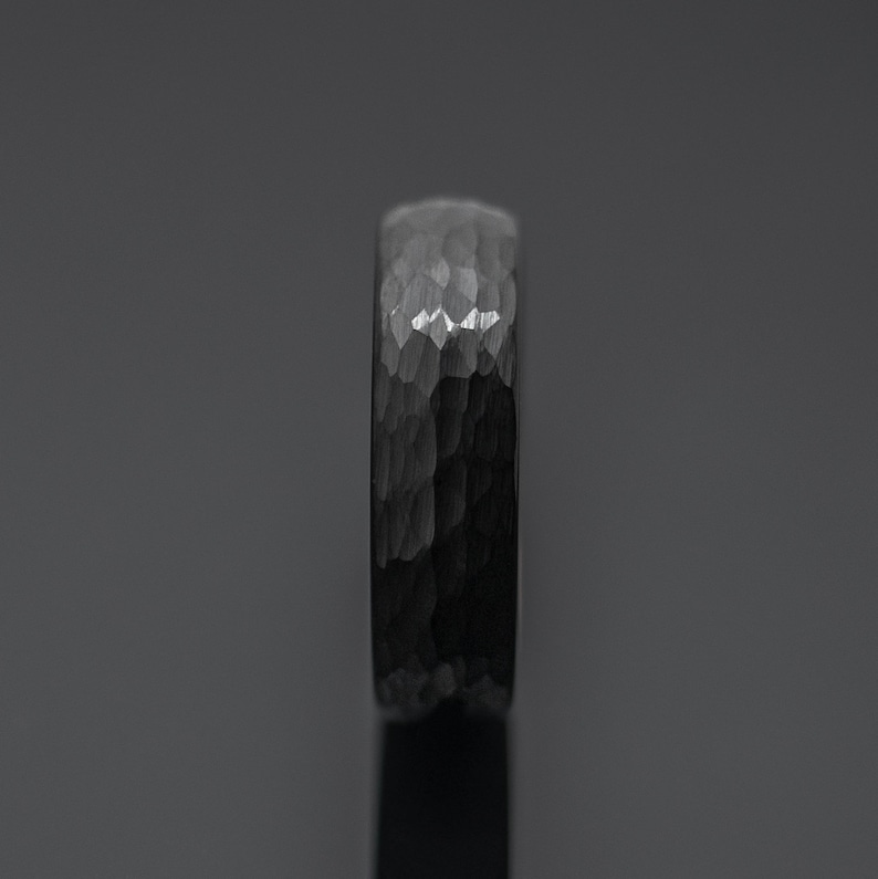 Op de afbeelding: Een donkergrijze ring met een gehamerde textuur. De ring heeft een bandbreedte van ongeveer 6 mm. Het oppervlak van de ring reflecteert licht, waardoor highlights en schaduwen ontstaan. De ring is een sieraad.